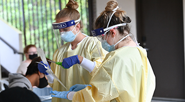 Two female UNF students in scrubs and face shields conducting COVID-19 testing on campus.