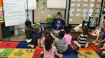 UNF preschool educator teaching students in a classroom. Everyone is wearing face masks while sitting on the floor.