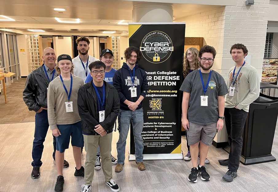 The Osprey Security student team posing in front of a Cyber Defense Competition banner