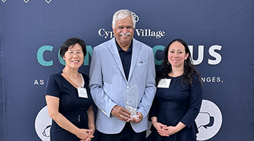 Dr. Mei Zhao, Dr. Shyam Paryani, and Sara Ninya posing in front of a Cypress Village canvas with their award