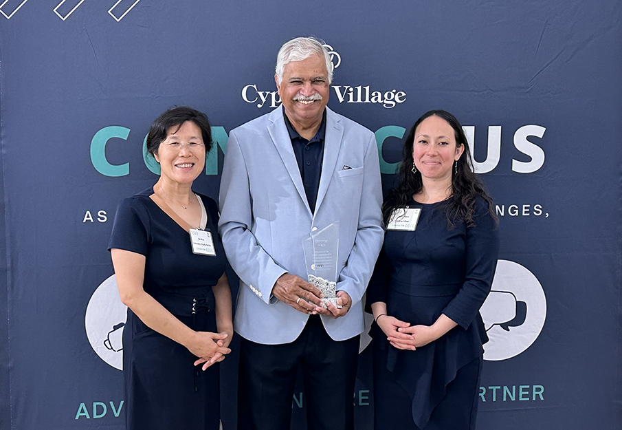 Dr. Mei Zhao, Dr. Shyam Paryani, and Sara Ninya posing in front of a Cypress Village canvas with their award