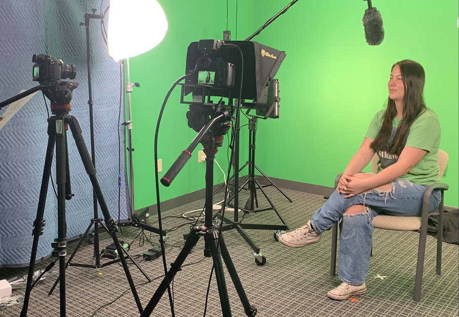 Student research assistant, Sarah Shiell, sitting in front of a green screen with a microphone and camera pointed at her