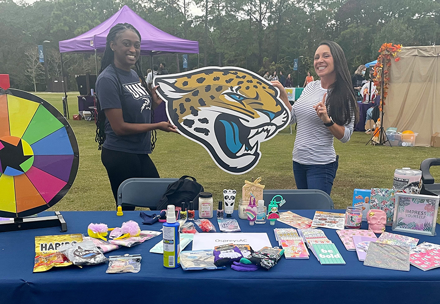Two ESG members holding up a team Jaguar head at a tabling event, standing behind a table that has the words "Eta Sigma Gamma, Health Education Honorary Society' written on the fabric