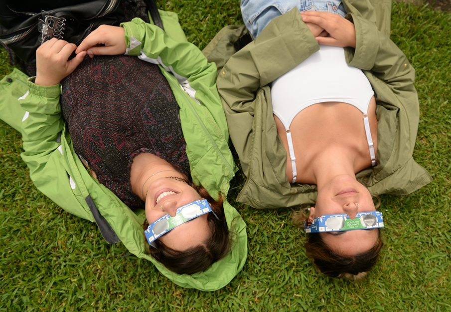 Aerial view of two students laying on the ground looking up at eclipse with special glasses