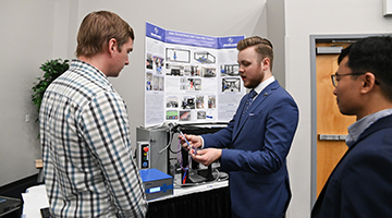 Engineering student presenting at Innovation Day in front of a poster board, holding up his invention