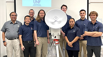 UNF student team and instructor Dr. Nirmal Patel that is participating in two NASA research projects posing in a group photo next to a satellite
