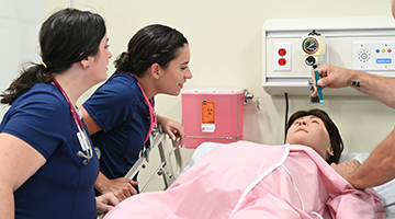 Two nursing students observing a professor demonstrate a wellness check