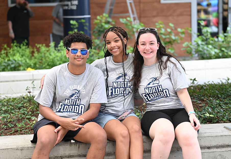 Three students wearing UNF t-shirts sitting together smiling at the camera at an event
