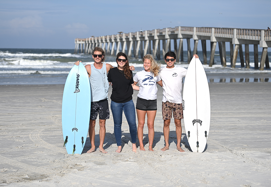 Four members of the UNF surf team posing together beside a surf board