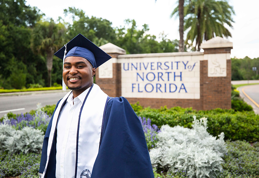 Tavian Cox, summer graduate, posing in front of the UNF sign in his cap and gown