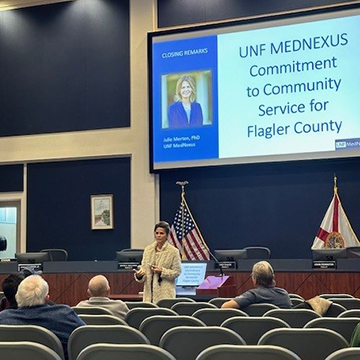 Dr. Julie Merten presenting in front of a screen that reads 'UNF MEDNEXUS Commitment to Community Service for Flagler County'