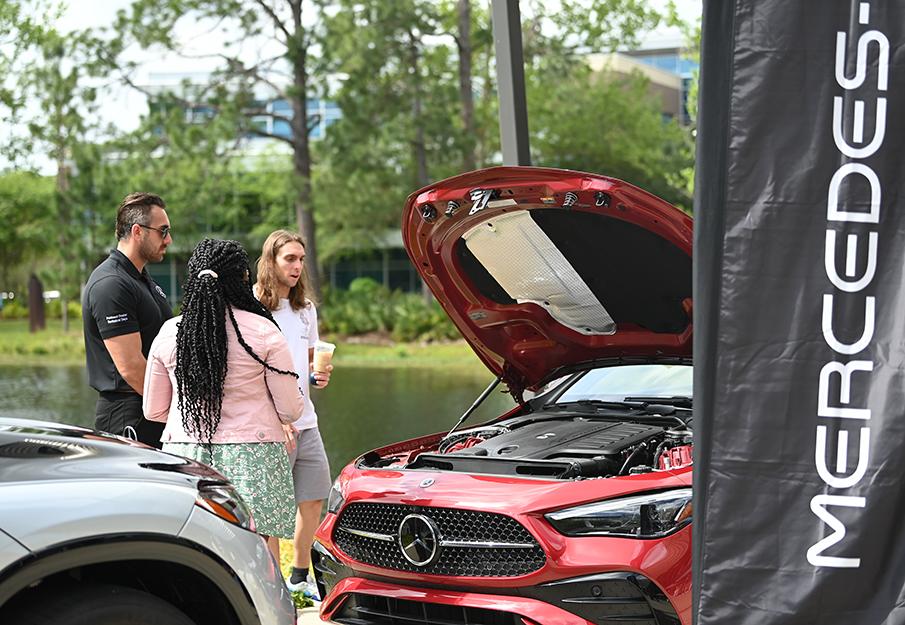 Two students speaking with a Mercedes employee while looking at a red Mercedes