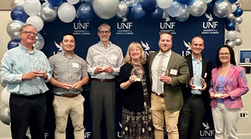 David Bauerlein, Chris Hong, Carla Miller, Nate Monroe, Mark Wood and Anna Brosche standing in front of a UNF backdrop holding their awards