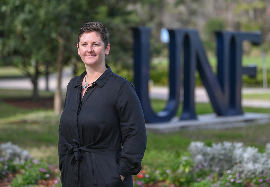 Dr. Sarah Mattice wearing all black standing on UNF campus with large UNF letters in the background
