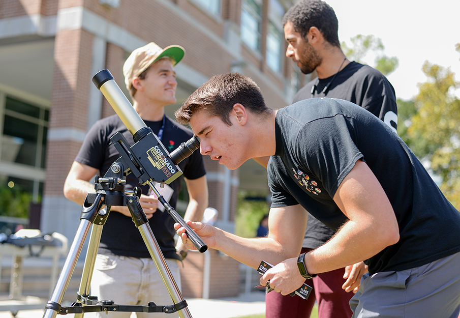 Two physics students talking while a third looks through a telescope