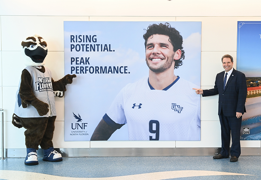 University of North Florida President Moez Limayem and mascot Ozzie pointing at a new sign that reads 'Rising Potential. Peak Performance.'