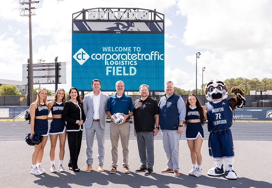 Three UNF cheerleaders, Dr. Angela Falconetti, Ozzie and Corporate Traffic and Athletics staff in front of a digital sign that reads WELCOME TO CORPORATE TRAFFIC LOGISTICS FIELD