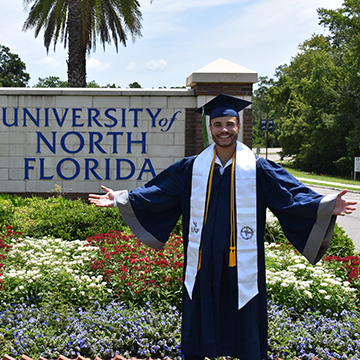 Michael Carrero in cap and gown posing in front of UNF sign