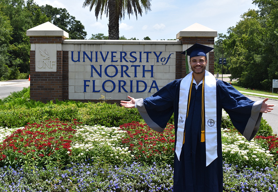 Michael Carrero in cap and gown posing in front of UNF sign