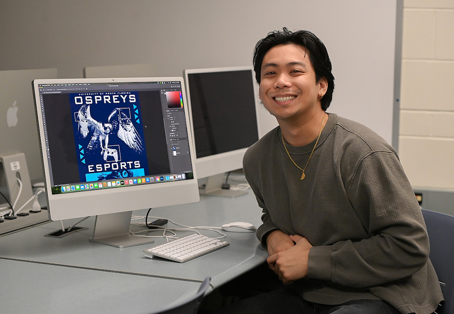 Matt Ducusin posing in front of his design; an osprey carrying a game controller with a blue background with the words University of North Florida Ospreys ESports