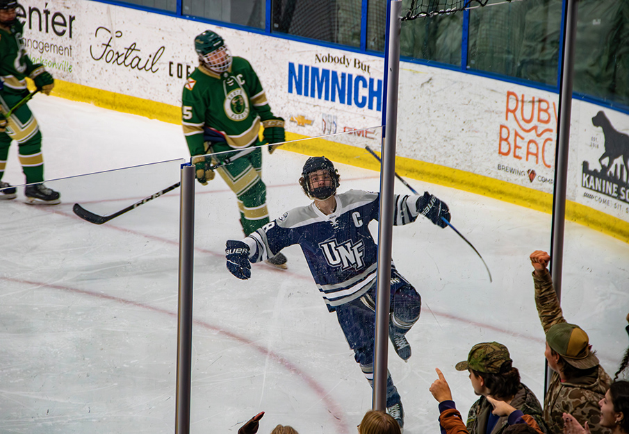 UNF Hockey player in mid 'swoop' on ice followed by two defeated St. John Fisher University players while crowd cheers