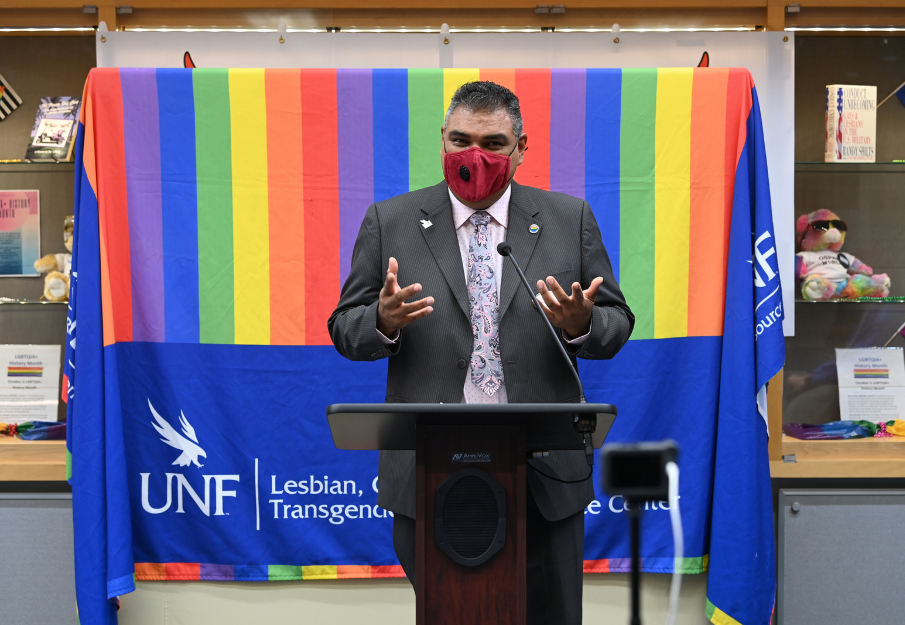 Director of the LGBTQ Center speaking at a podium with a rainbow banner behind him