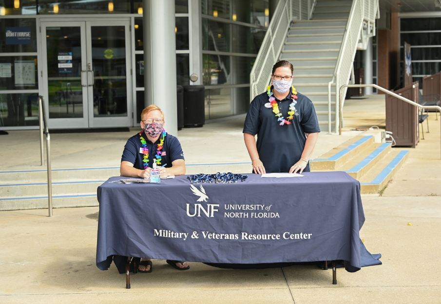 Two people sitting behind a militarys and veterans resource center table waving at the camera