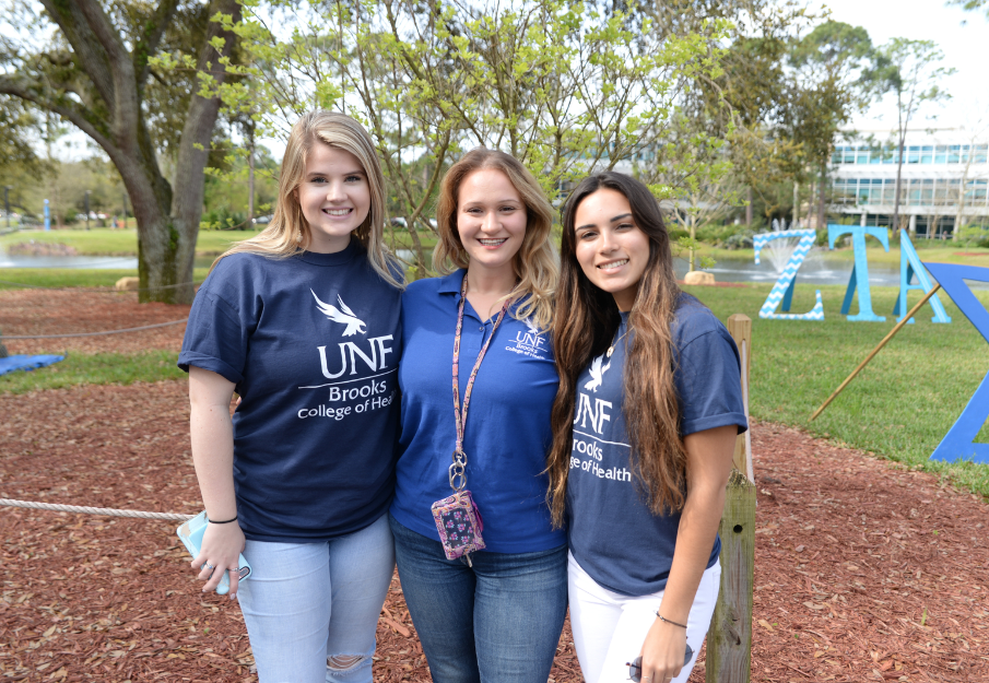 Three female students standing on campus with Brooks College of Health t-shirts