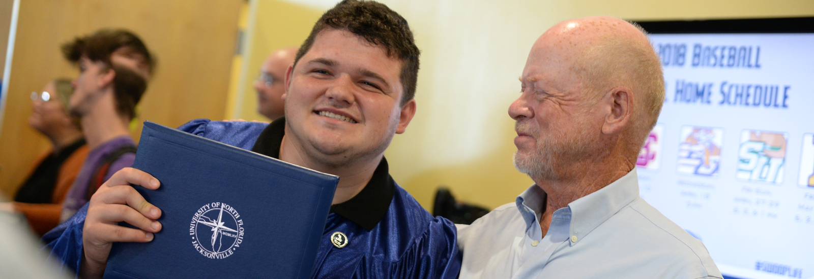 Male student in a UNF graduation gown holding a UNF diploma and standing by his dad