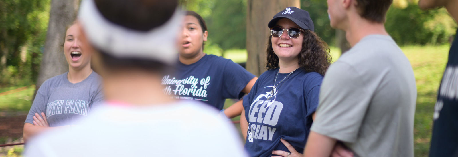 Female in sunglasses, wearing a UNF hat and t-shirt, with several other students out of focus around her