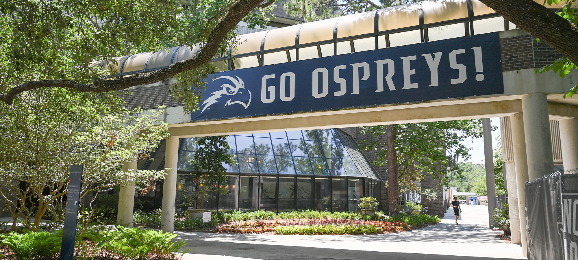 Nature surrounding a walkway UNF bridge with a sign saying Go Ospreys!