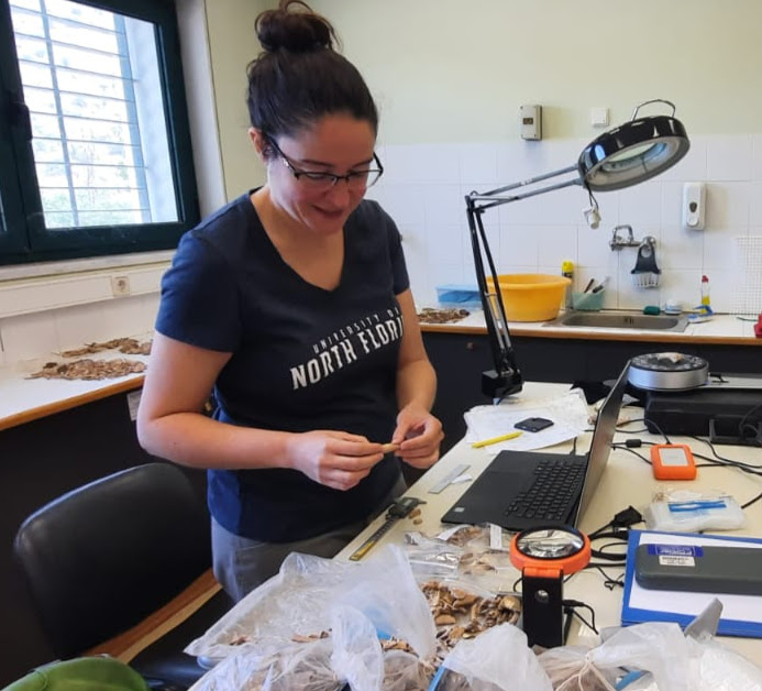 Jackie sorting through bone fragments in her lab, she is surrounded by lab equipment and her hair is in a bun. 