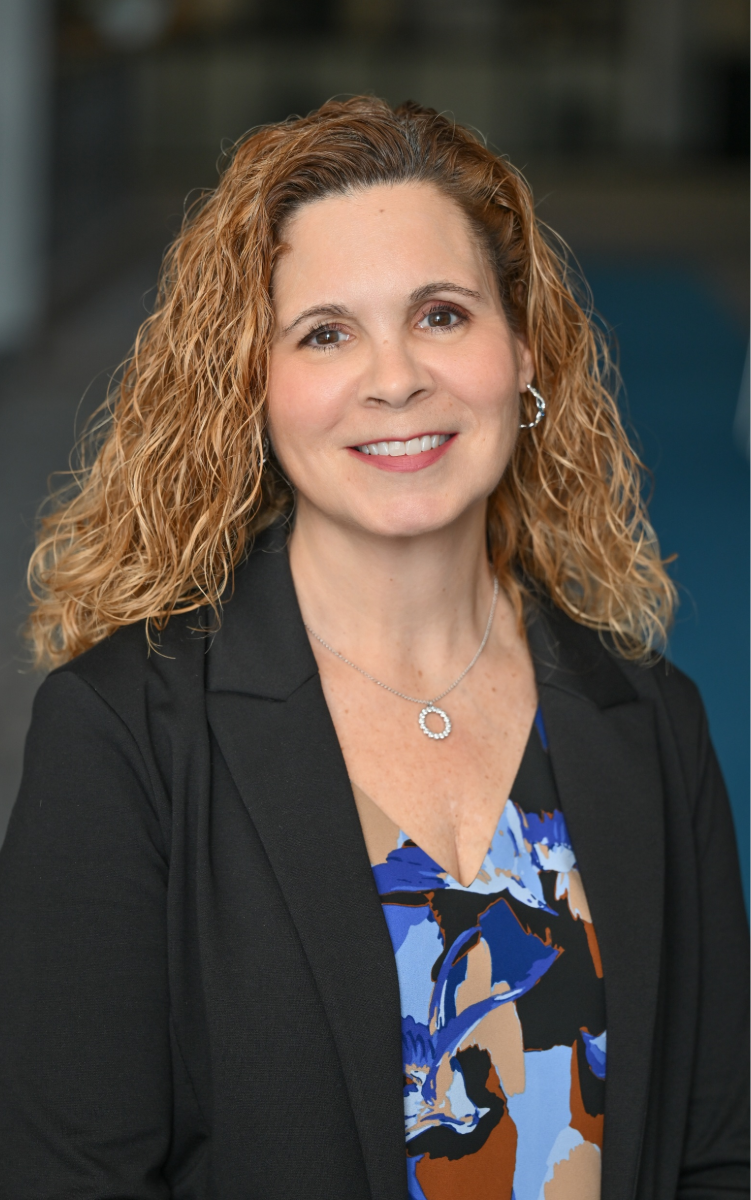 Headshot of Judy smiling at the camera. She is wearing a blue patterned shirt, black blazer, and silver jewelry. 