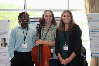 August Researcher's of the Month, Megan Herrman, Khalil Payne, and Grace Raper holding a cello and standing in front of their research poster