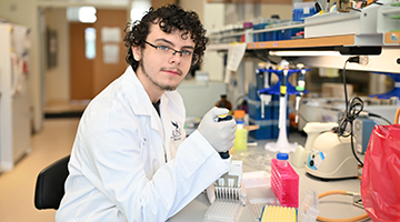 Cameron is in a lab coat and is using lab equipment. He has dark curly hair and glasses.