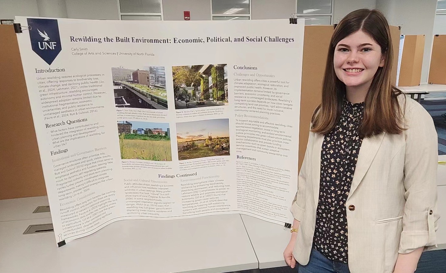 Carly Smith standing in front of her research poster and smiling. She is wearing a tan blazer, dark floral shirt, and jeans.