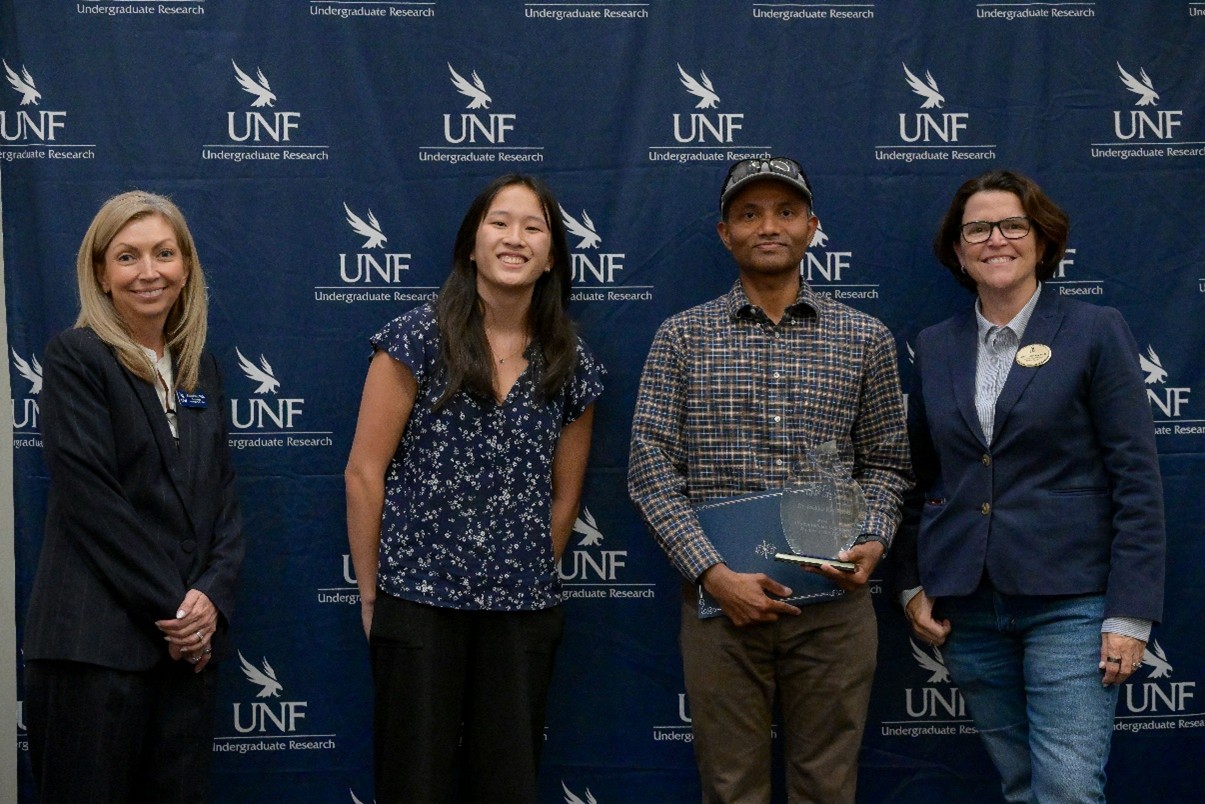 Dr. Kahanda is standing in front of a blue photo back drop with the UNF OUR logo on it. He is wearing a gray hat with sunglasses on his head, and a blue and tan checkered shirt. He is holding a certificate and award plaque alongside 3 women.
