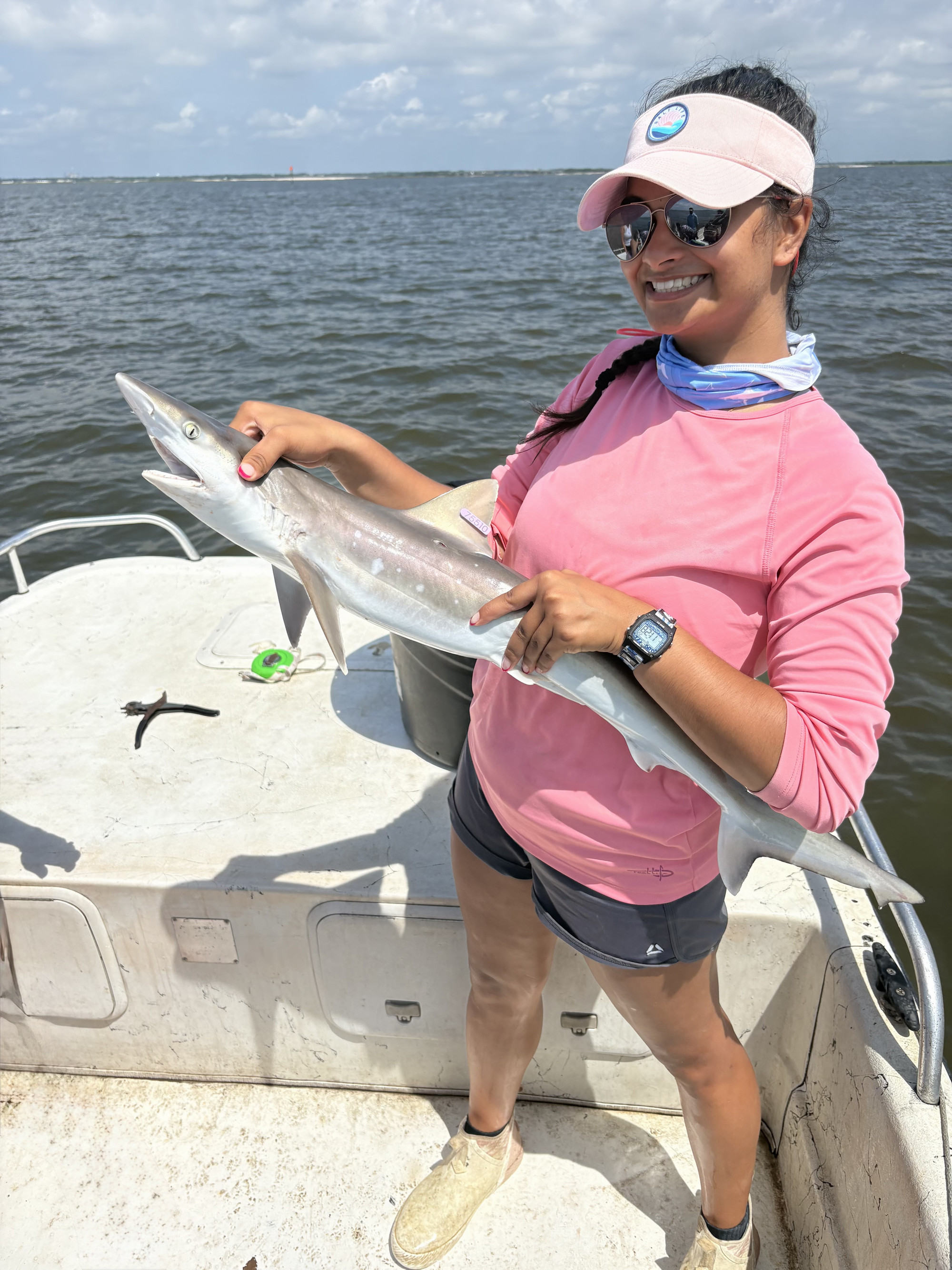Jordyn standing on a boat and holding a small shark. She is wearing a pink shirt and is smiling. 