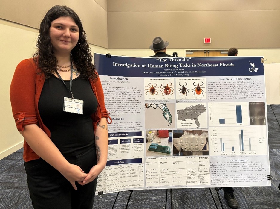 Kiersten is standing in front of her research poster and smiling. She is wearing a black shirt and red cardigan.