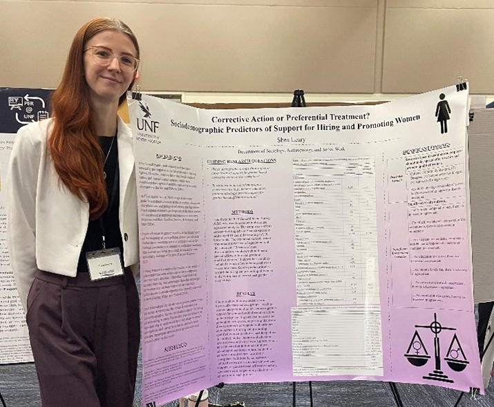 Shea is standing in front of her research poster and smiling. She has red hair and is wearing glasses, a white blazer, gray pants, and a black shirt.