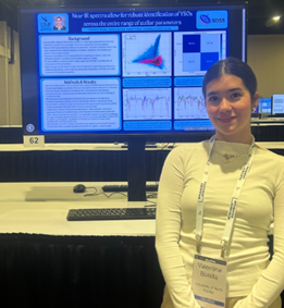 Valentina standing in front of an electronic poster with her hands crossed. Her hair is tied back and she is wearing a white shirt and a participant event lanyard.
