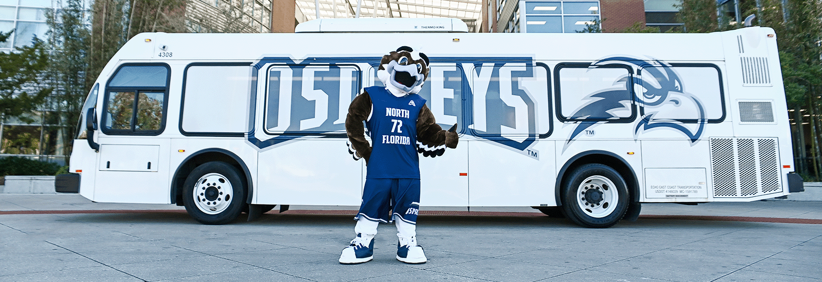 Ozzie is standing in front of Osprey Connector shuttle that is parked in Lot 4 and in front of the Student Union buildings