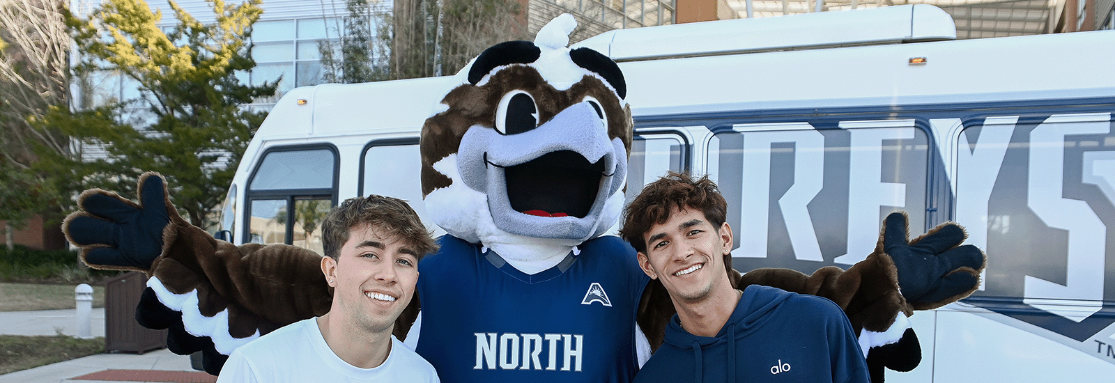 Two students posing with Ozzie, UNF's mascot, in front of a campus shuttle