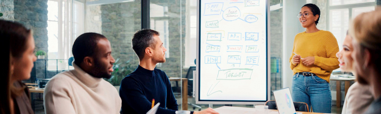 Project management team meeting around a table with a woman standing near a white board