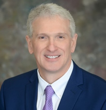 A smiling headshot of Vice President Jay Coleman. VP Coleman is wearing a navy blue suit with a purple tie with white polka dots.