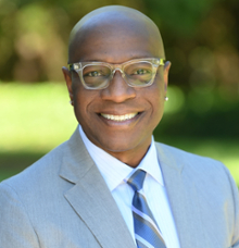 A smiling headshot of Vice President Richmond Wynn. VP Wynn is wearing a navy blue suit with a light blue tie. VP Wynn is wearing black glasses. 