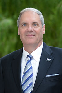 A smiling headshot of President Szymanski wearing a navy blue suit with a blue and grey stripped tie. 