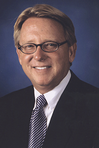 A smiling headshot of President Delaney wearing a black suit with a black and white stripped tie as well as black glasses. 