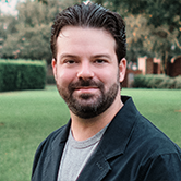 Head-and-shoulders outdoor portrait of a bearded man with dark hair, wearing a dark jacket over a gray shirt, with a blurred green lawn and trees in the background.