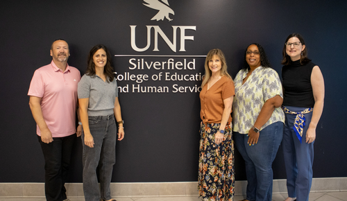Five people stand in front of a wall sign reading “UNF Silverfield College of Education and Human Services.”